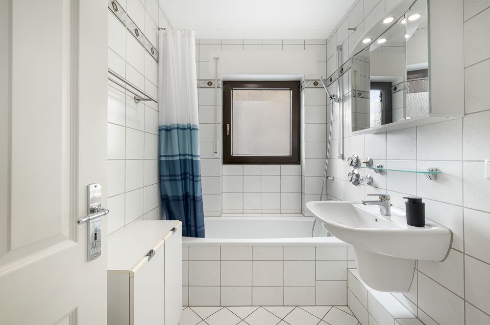 Helles Badezimmer mit Badewanne, Waschbecken und Fenster, weiße Fliesen an Wänden und Boden.