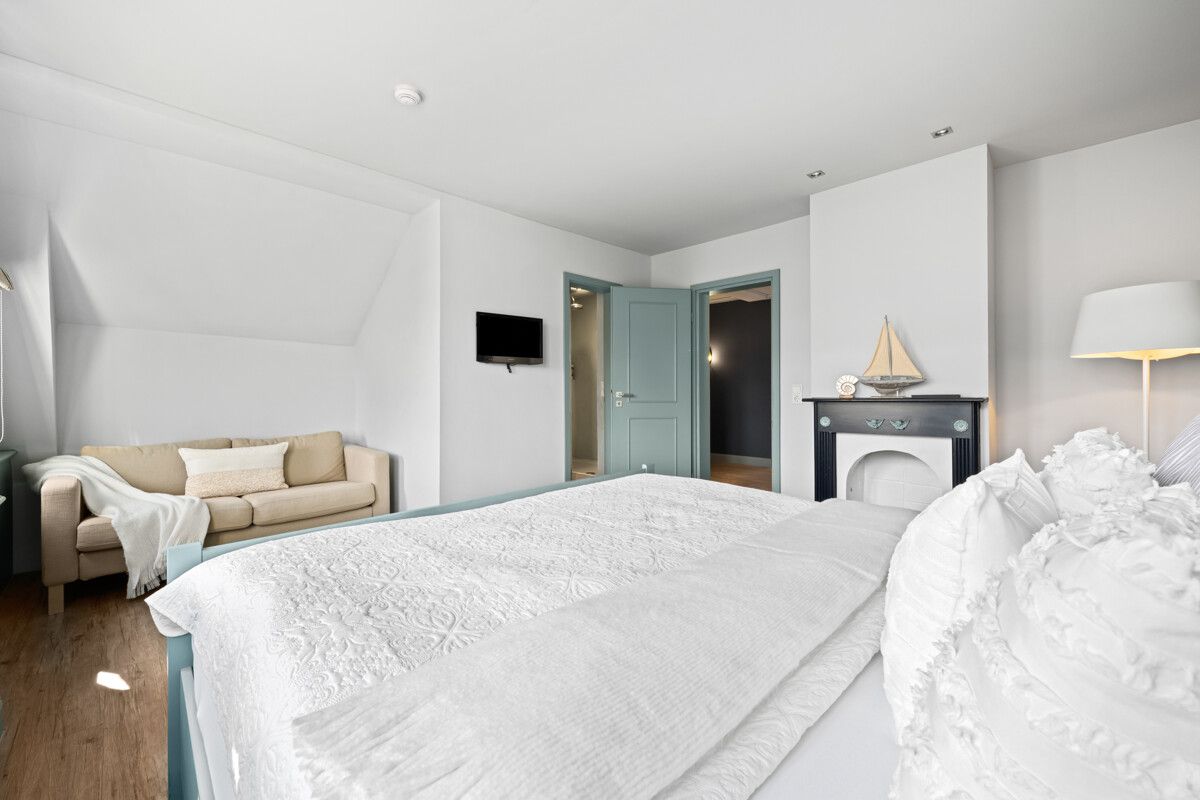 Helles Schlafzimmer mit Doppelbett, Sofa, Kamin, Stehlampe und Fernseher an der Wand.