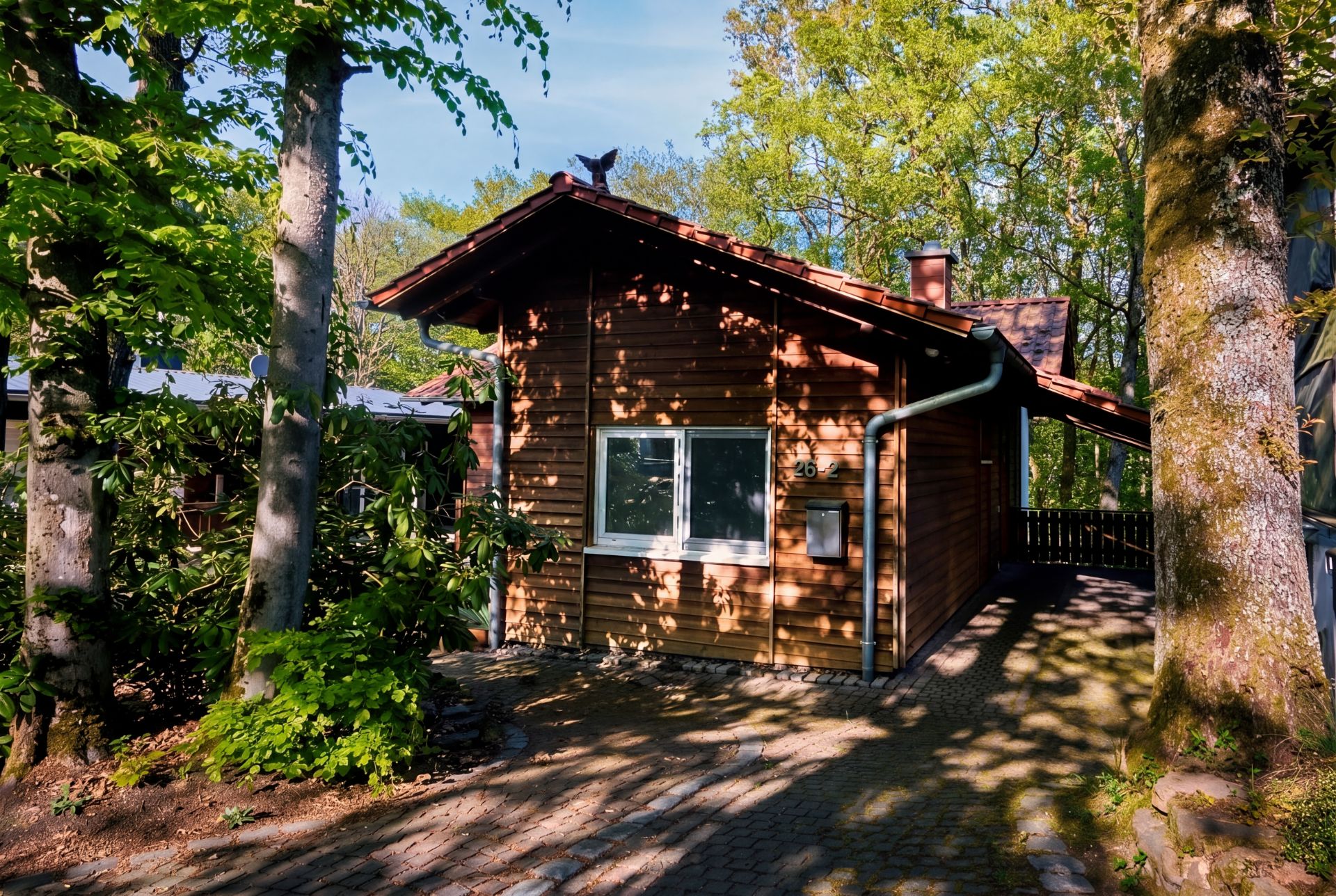 Kleines Holzhaus mit Fenster und gepflastertem Weg, umgeben von Bäumen und Pflanzen.