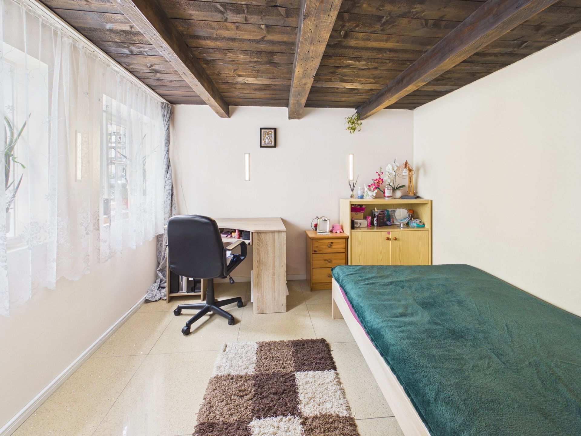 Helles Zimmer mit Holzbalkendecke, Bett, Schreibtisch mit Stuhl und Teppich auf Fliesenboden.