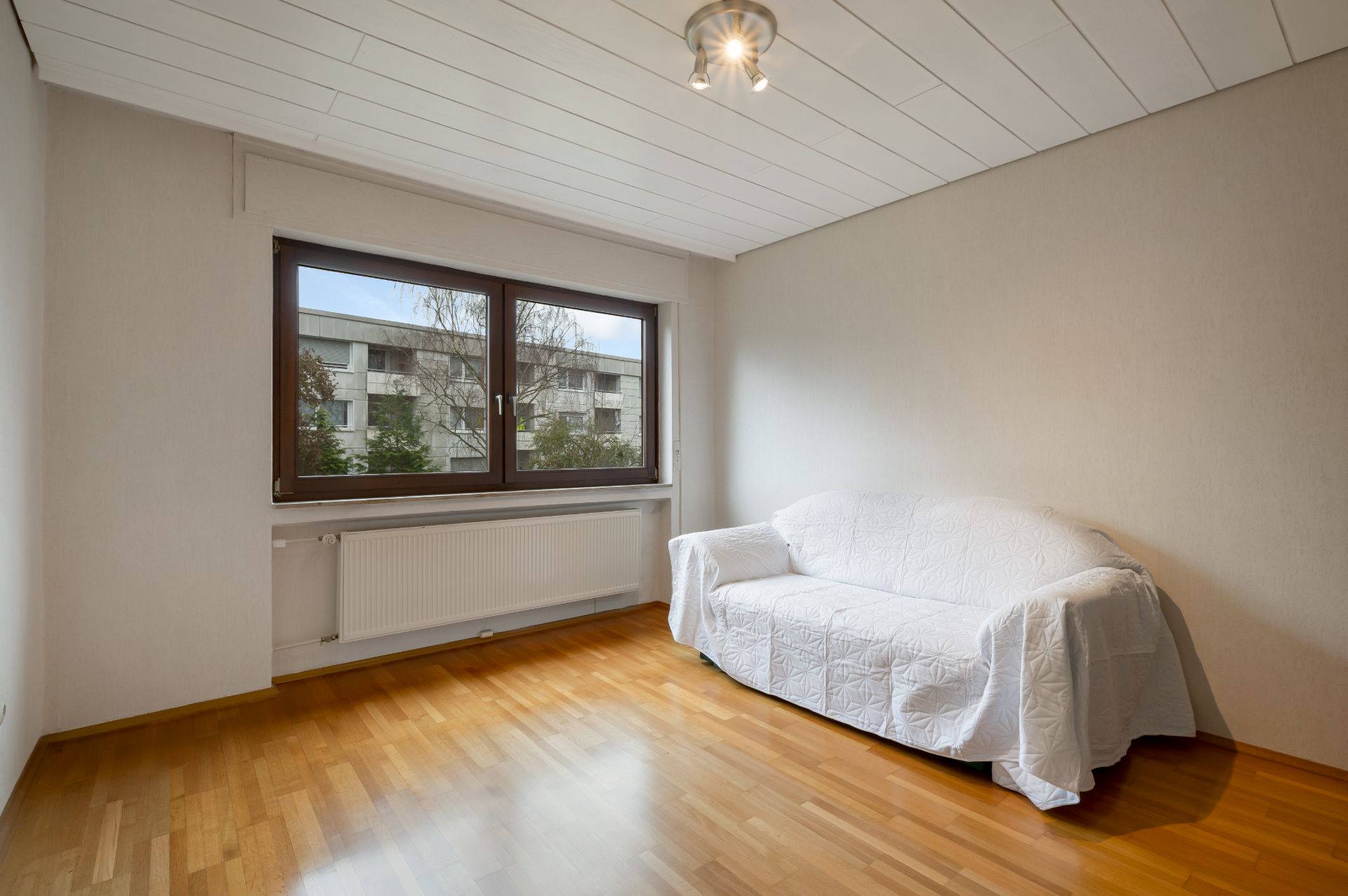 Helles Zimmer mit Parkettboden, einem Fenster und einem Sofa mit weißem Überwurf.