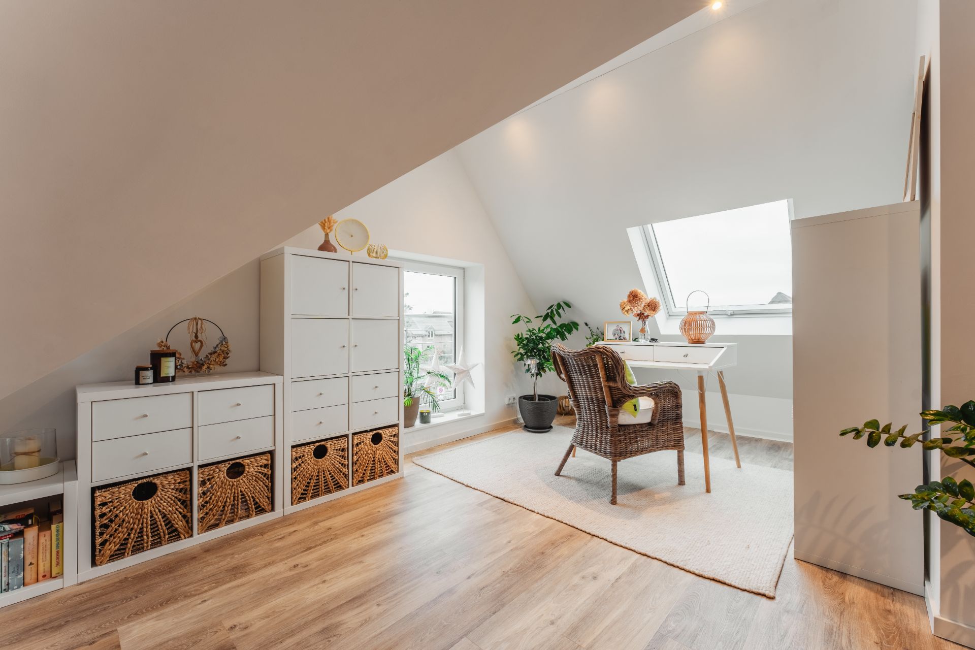 Helles Dachzimmer mit Schreibtisch, Korbstuhl, Pflanzen und weißen Regalen auf Holzfußboden.