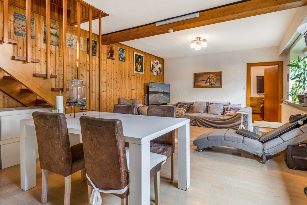 Wohnzimmer mit Holzwand, Sofa, Essbereich und offener Treppe, helle Einrichtung und Pflanzen am Fenster.