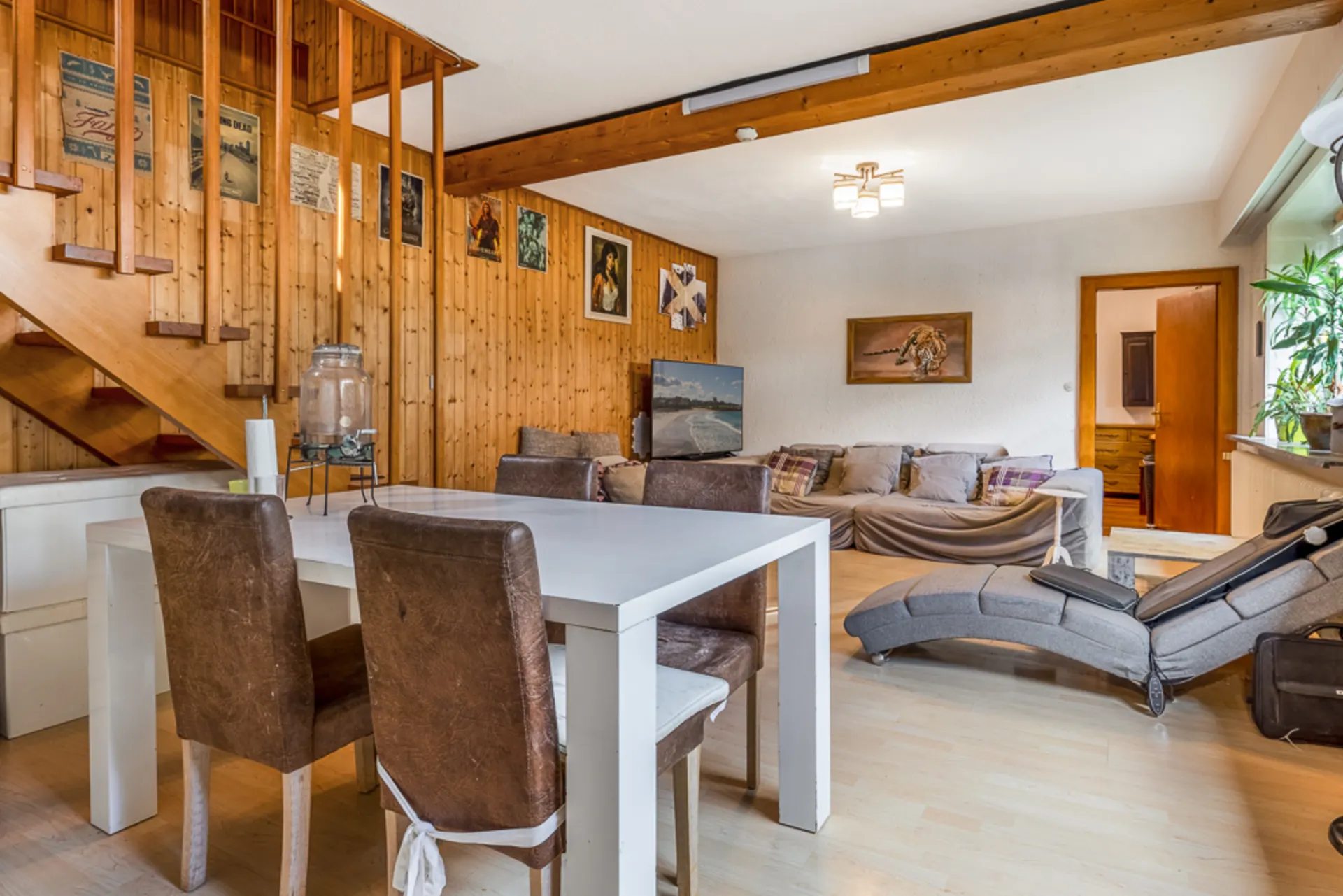 Wohnzimmer mit Holzwand, Sofa, Essbereich und offener Treppe, helle Einrichtung und Pflanzen am Fenster.