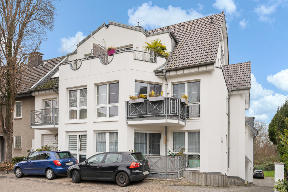 Modernes, weißes Wohnhaus mit Balkonen, großen Fenstern und mehreren geparkten Autos davor.