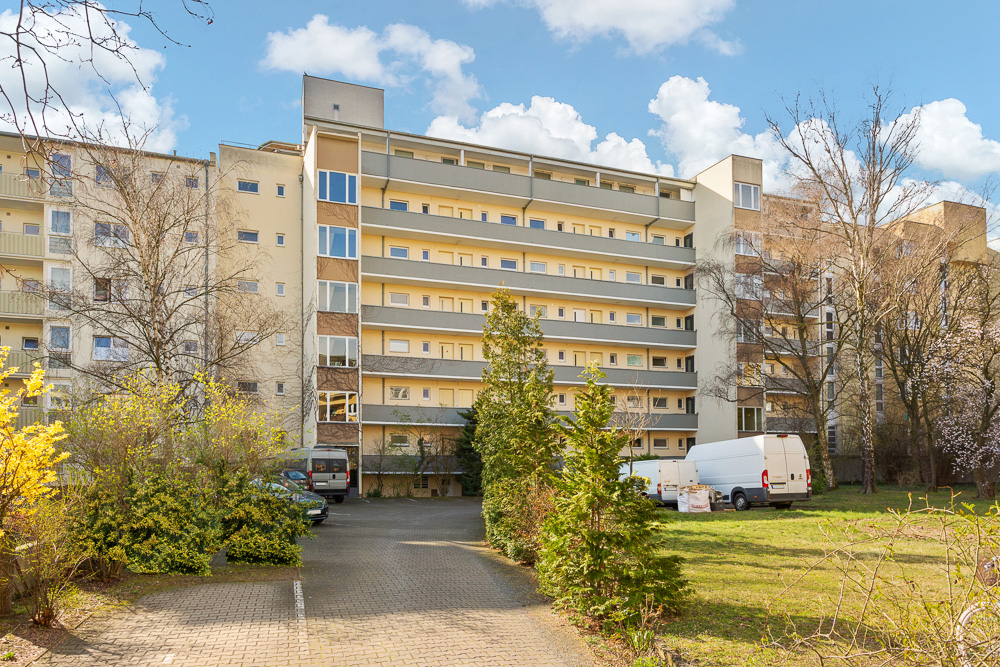 Mehrstöckiges Wohngebäude mit Balkonen, davor Bäume und geparkte Fahrzeuge.