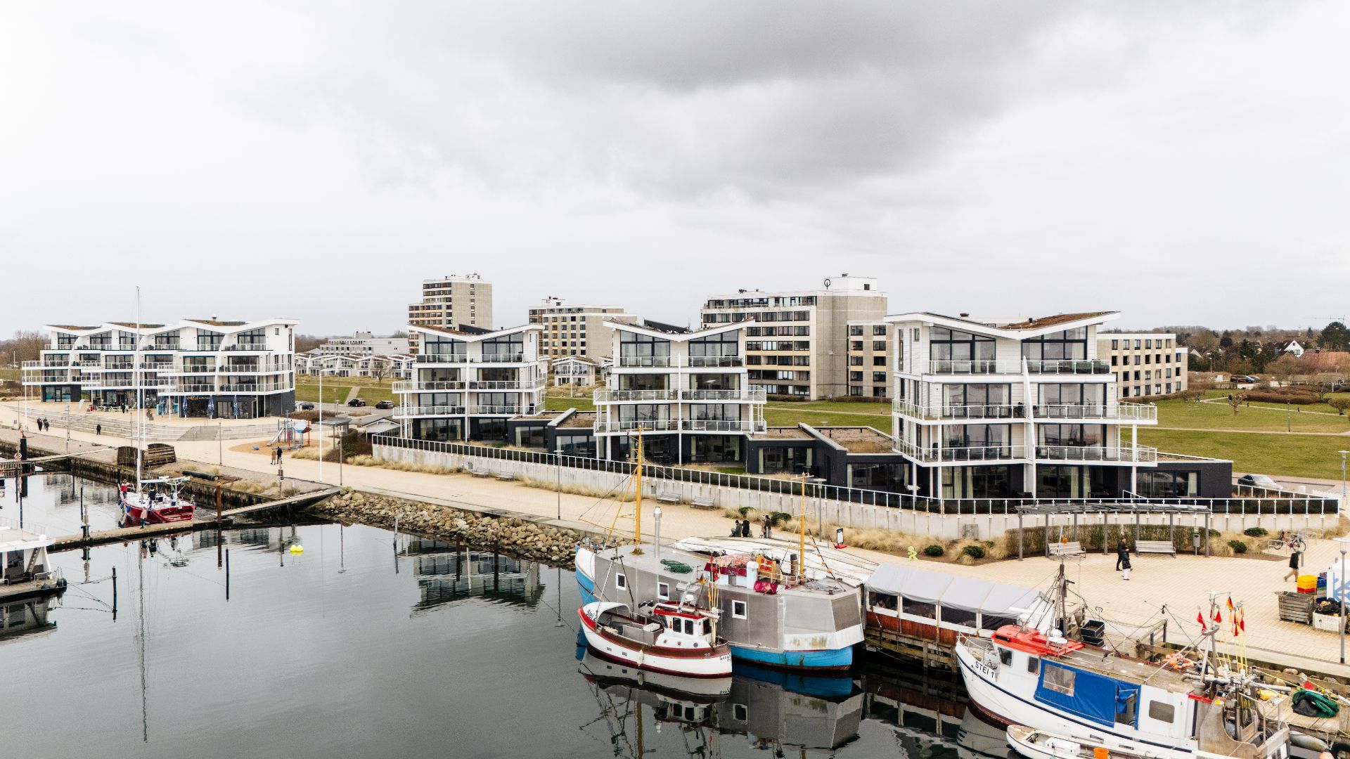 Moderne Wohngebäude am Wasser mit Booten im Vordergrund und bewölktem Himmel.