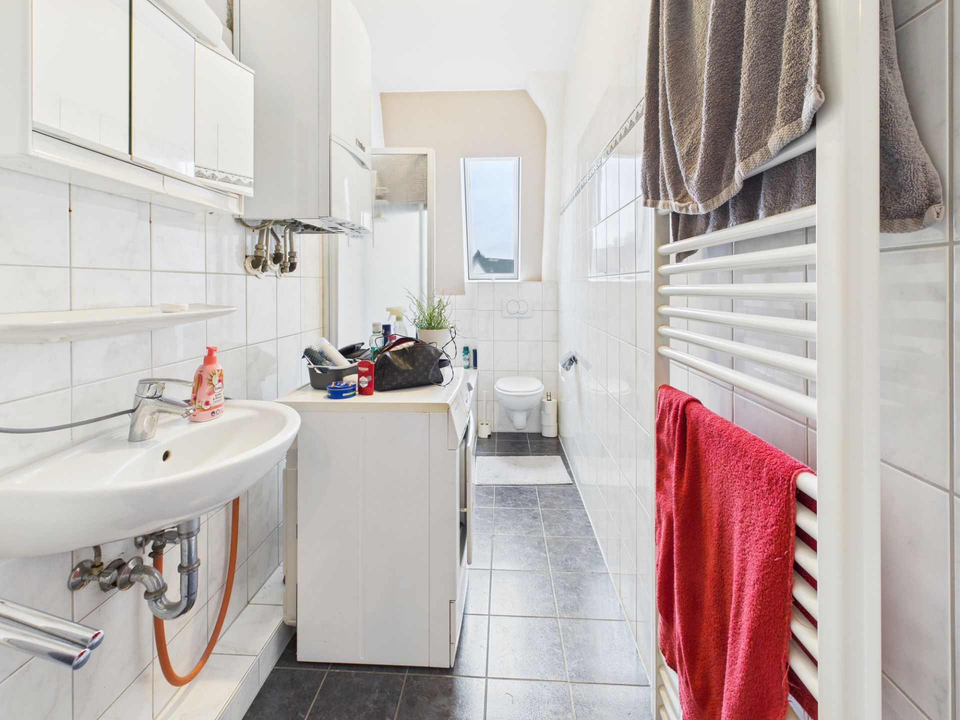 Schmales Badezimmer mit Waschbecken, Toilette, Handtuchheizkörper und Wandspiegel.