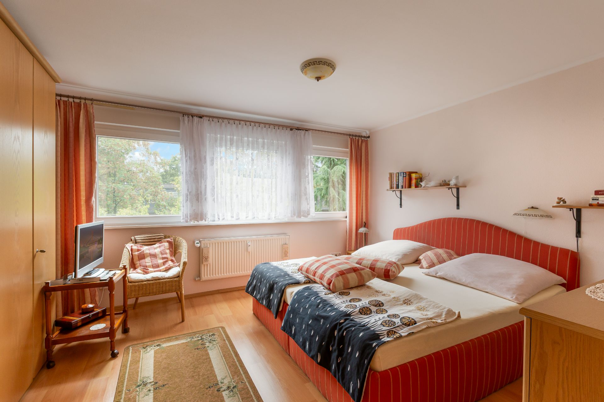 Helles Schlafzimmer mit Doppelbett, Sessel, Fernseher, Teppich und großen Fenstern.