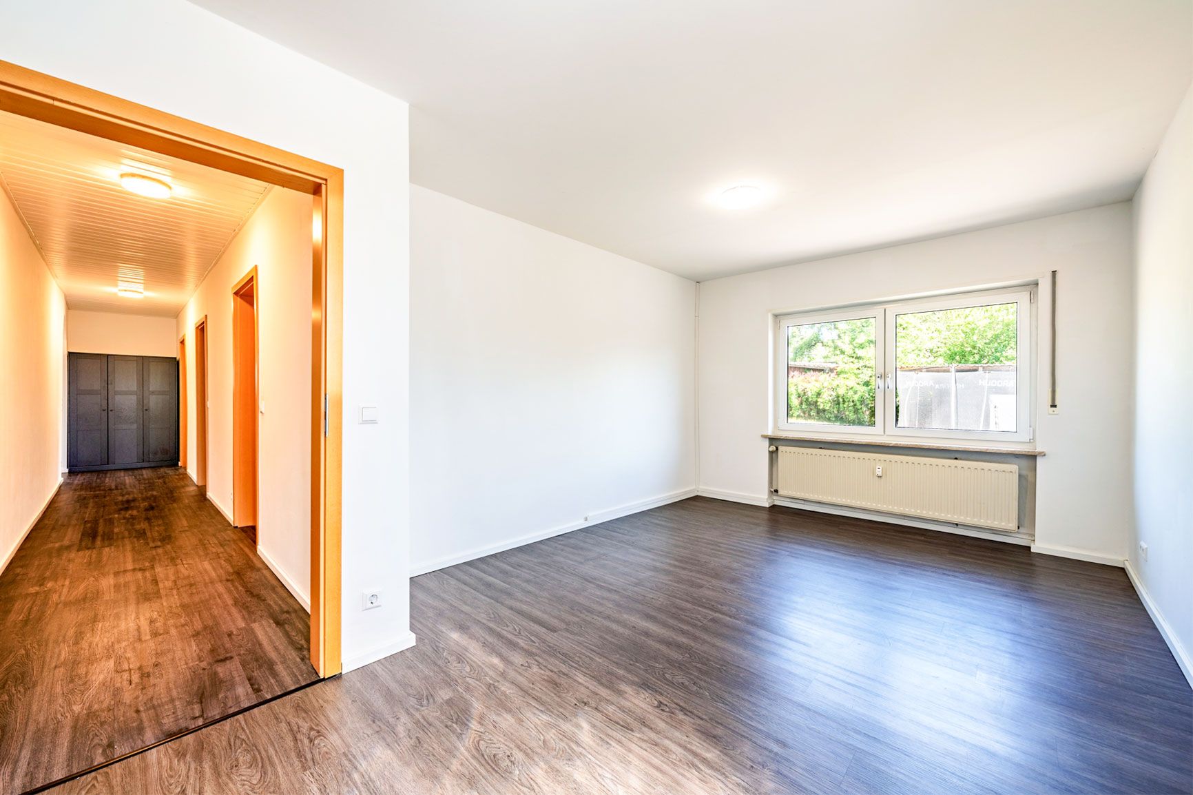 Helles Wohnzimmer mit großen Fenstern und dunklem Boden, Flur mit Türen im Hintergrund.