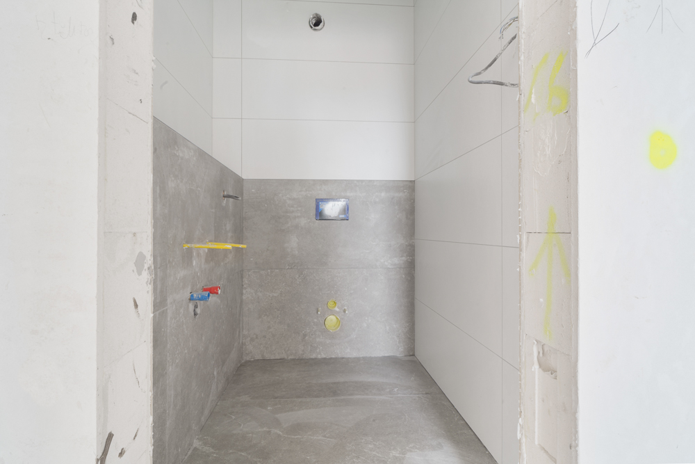 Unfertiges Badezimmer mit grau-weißen Fliesen und sichtbaren Sanitäranschlüssen.