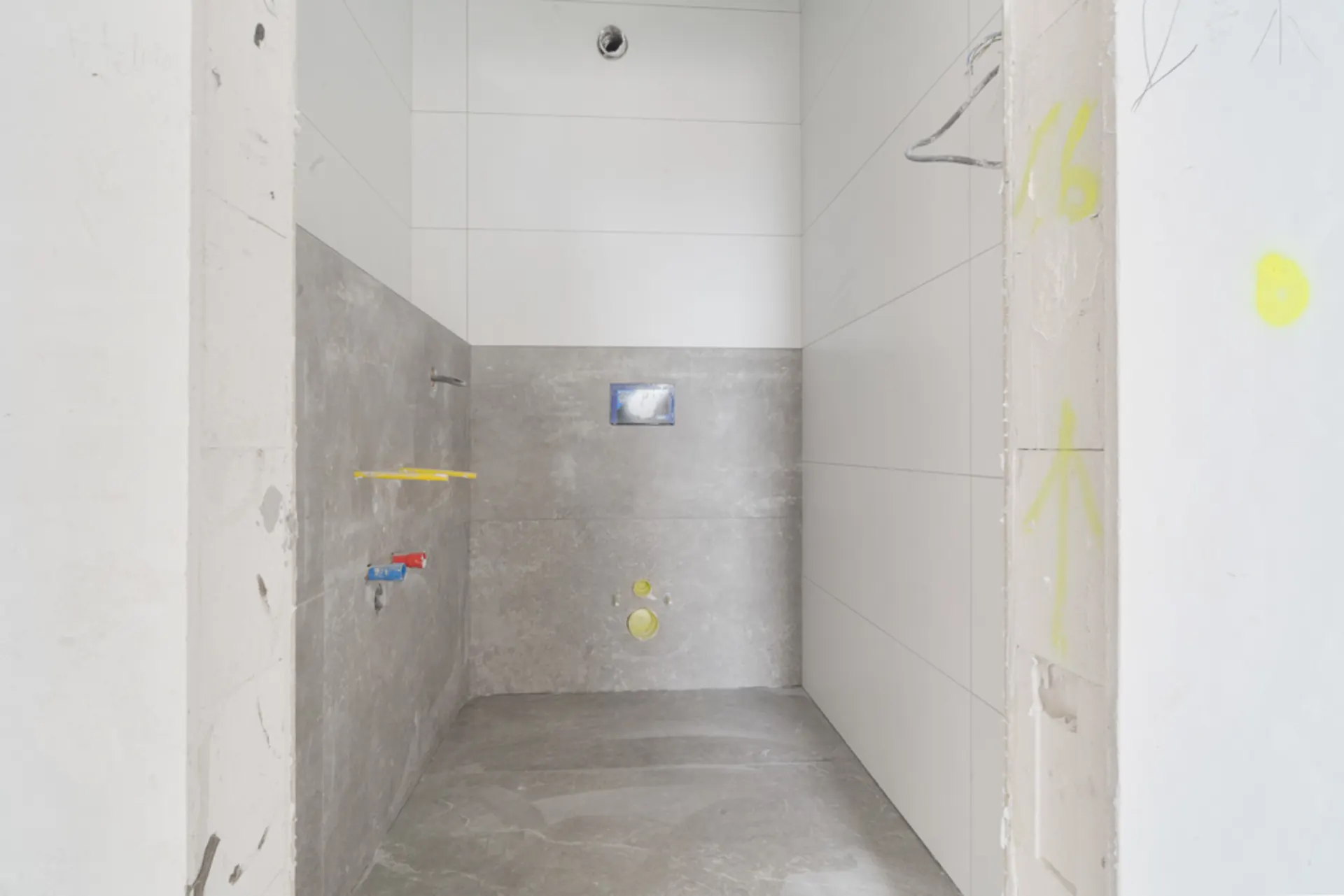 Unfertiges Badezimmer mit grau-weißen Fliesen und sichtbaren Sanitäranschlüssen.