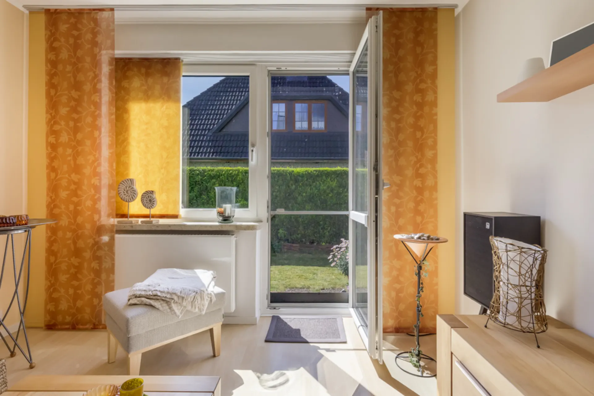 Helles Wohnzimmer mit offenem Fenster zur Terrasse, orange Vorhänge, Stuhl und Gartenausblick.
