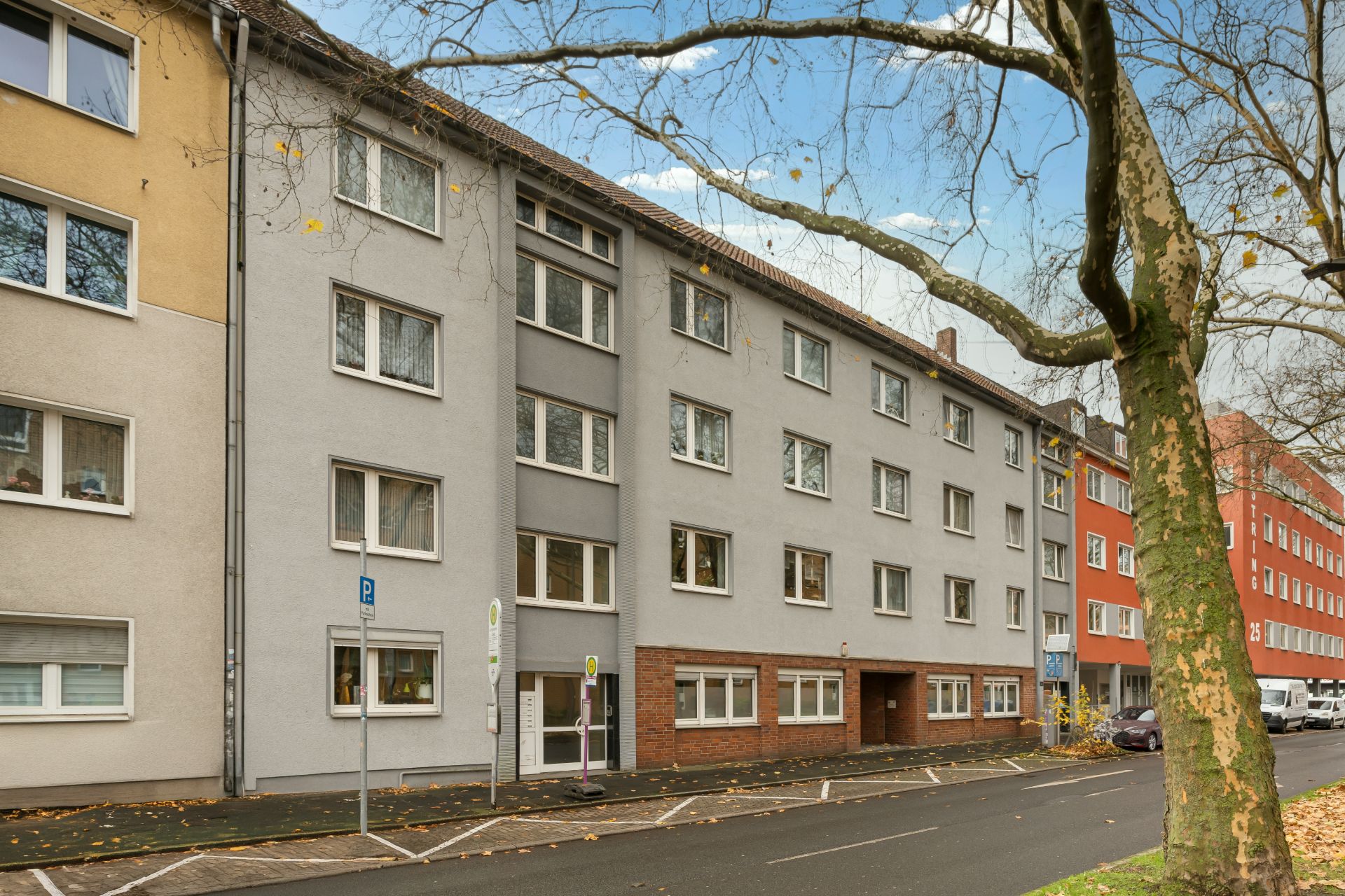 Mehrstöckiges Wohnhaus mit grauer Fassade, vielen Fenstern und Parkplätzen an der Straße.