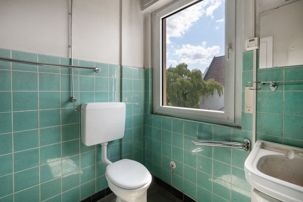 Kleines Bad mit grünen Fliesen, Toilette, Waschbecken und Fenster mit Blick nach draußen.