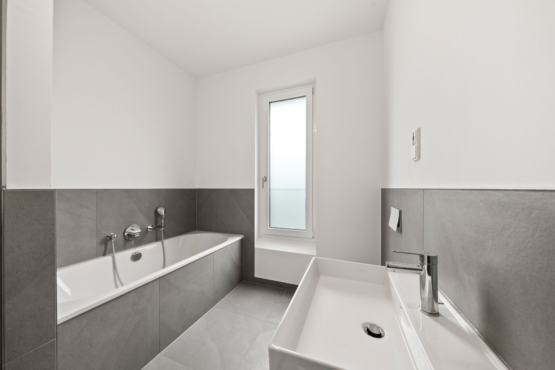 Modernes Badezimmer mit Badewanne, Waschbecken und Fenster, graue Fliesen an Wand und Boden.