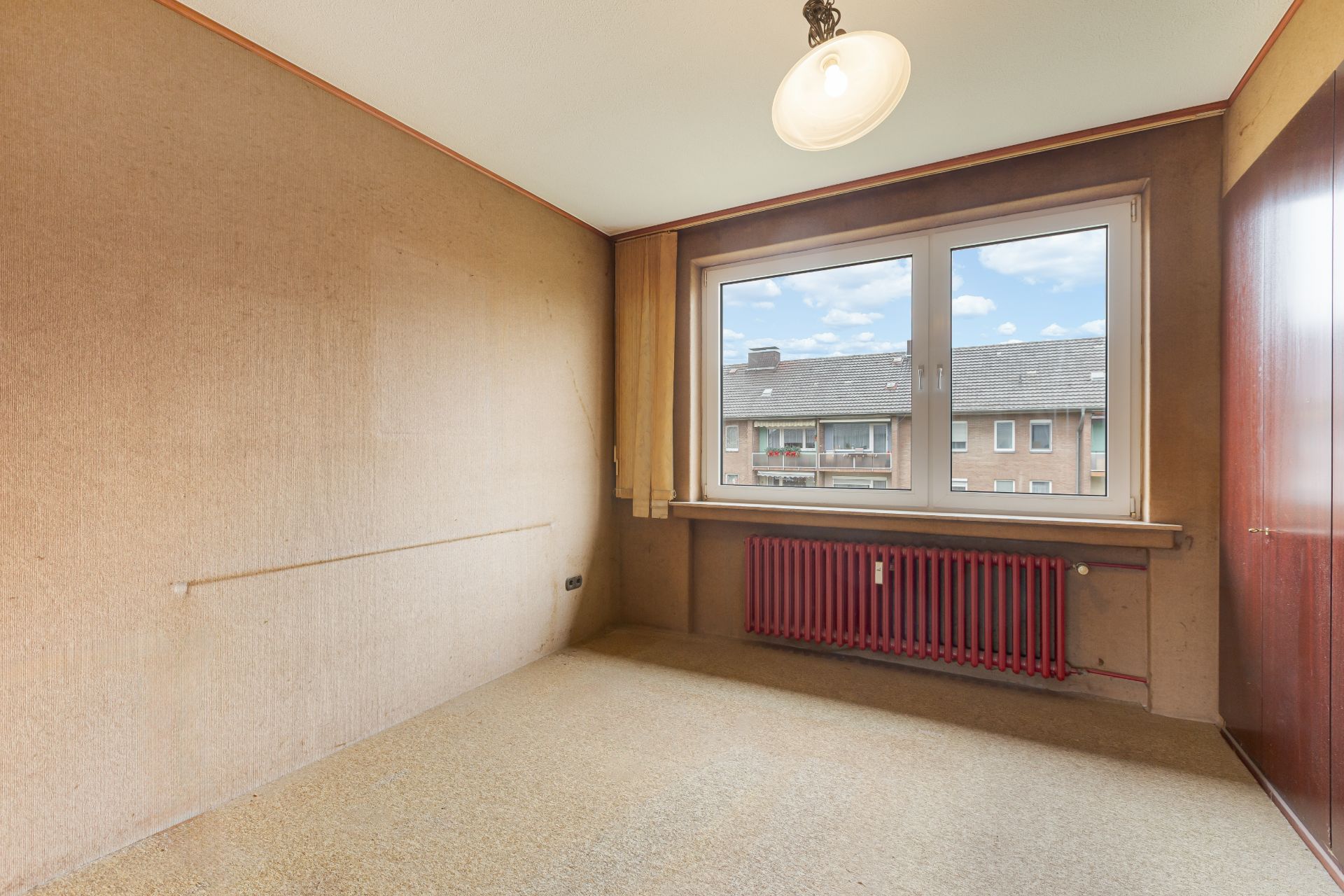 Leeres Zimmer mit Teppichboden, großem Fenster und rotem Heizkörper unter dem Fenster.