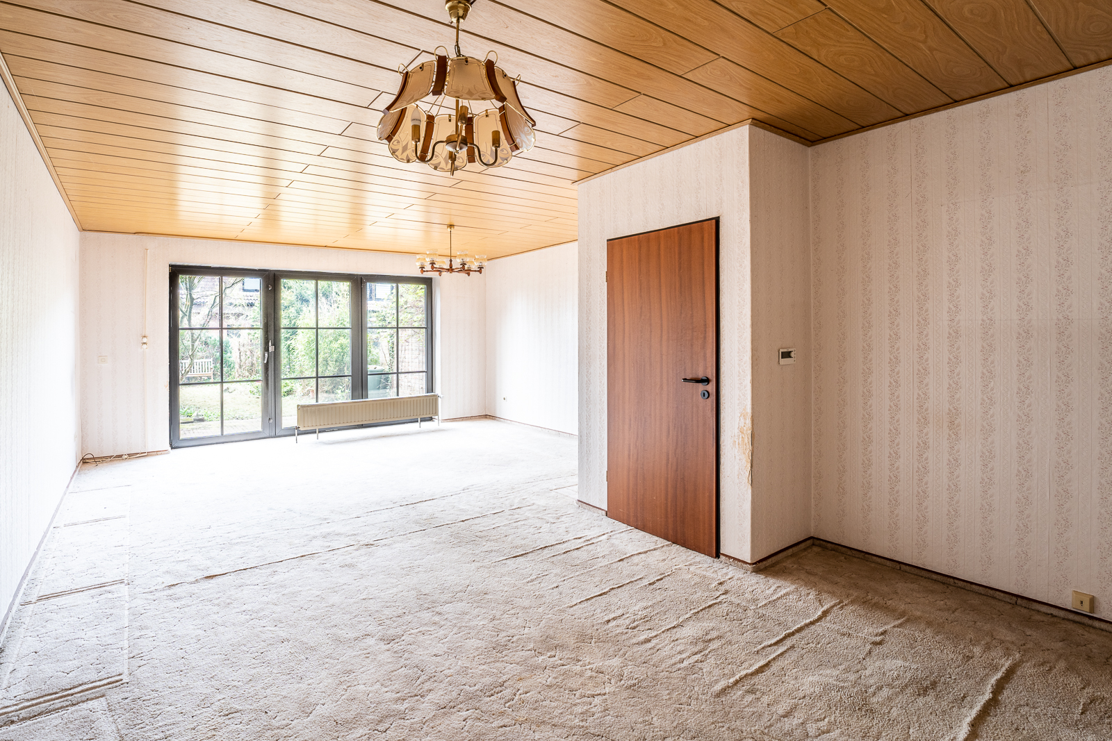 Leeres Wohnzimmer mit Holztür, Teppichboden, Holzdecke und großen Fenstern zum Garten.