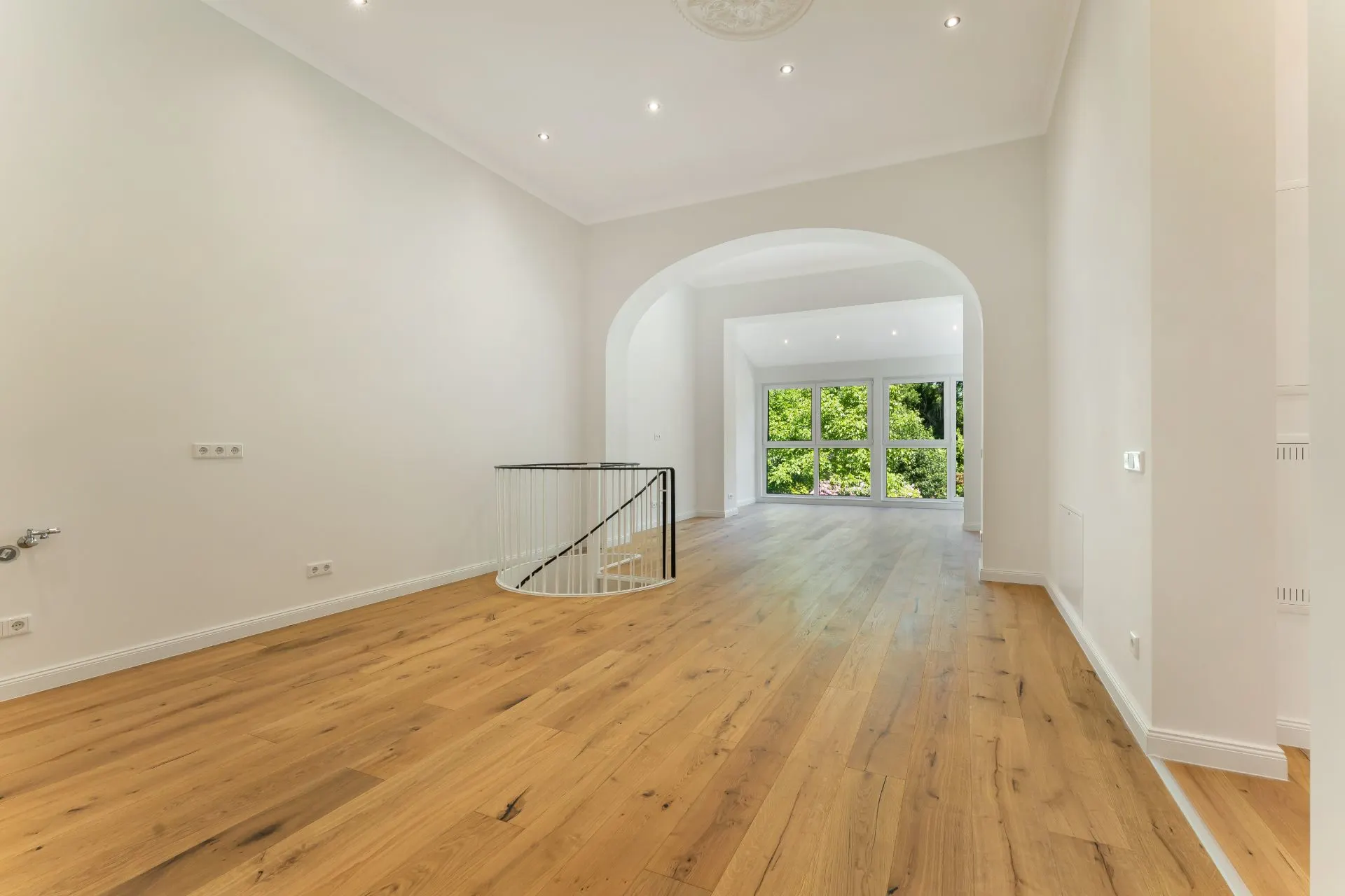 Heller Raum mit Holzboden, großen Fenstern und Treppe mit schwarzem Geländer.