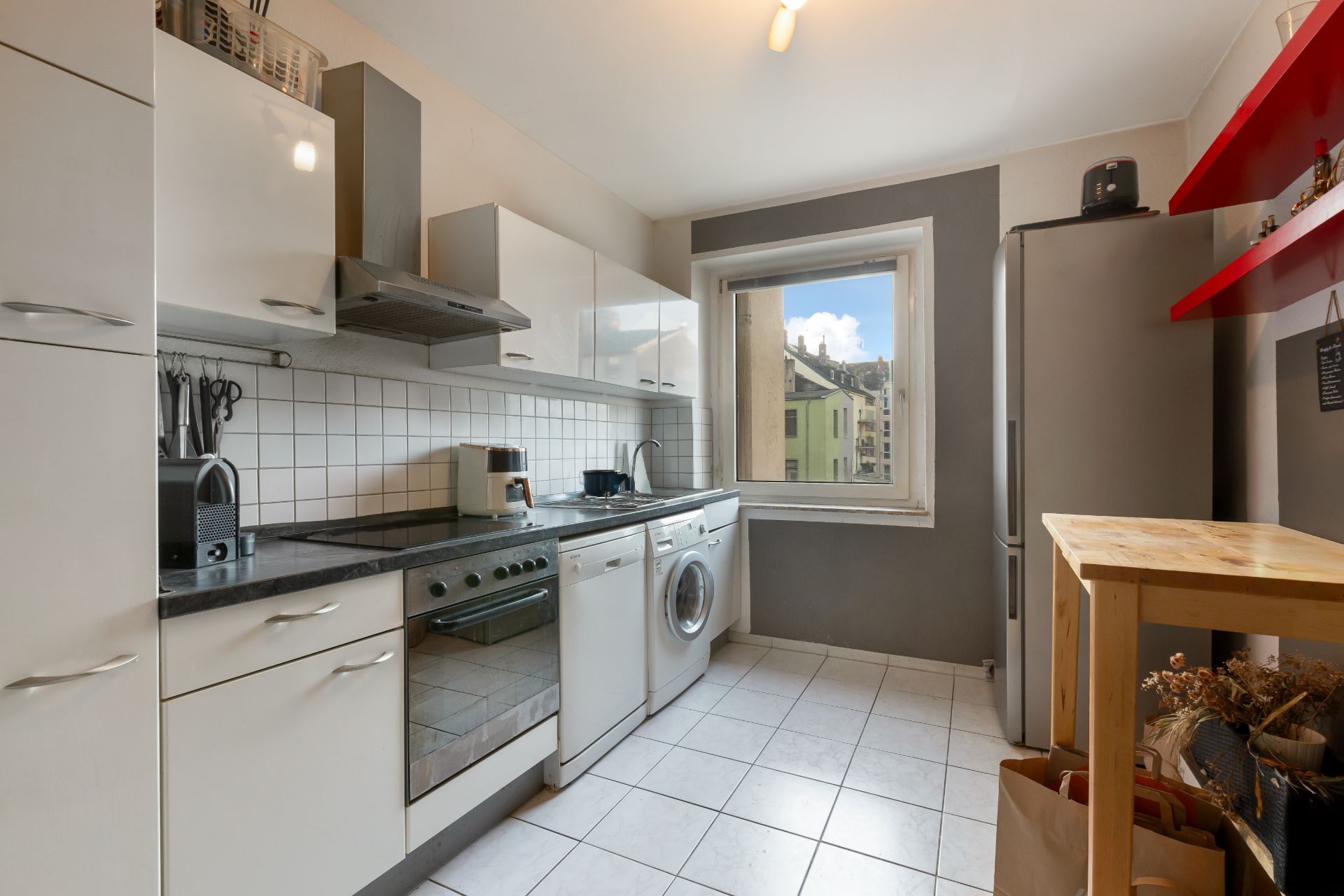 Helle Küche mit weißen Schränken, Elektrogeräten, Fenster und Holztisch.