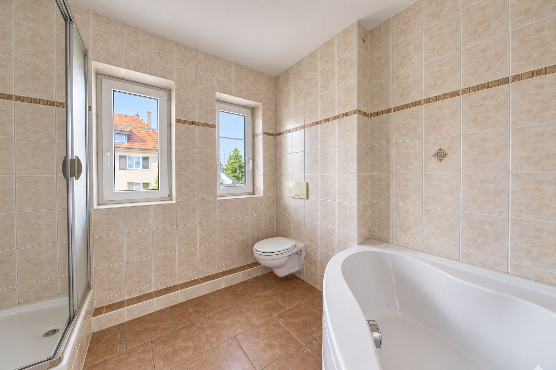 Helles Badezimmer mit Badewanne, Dusche, Toilette und zwei Fenstern mit Blick nach draußen.