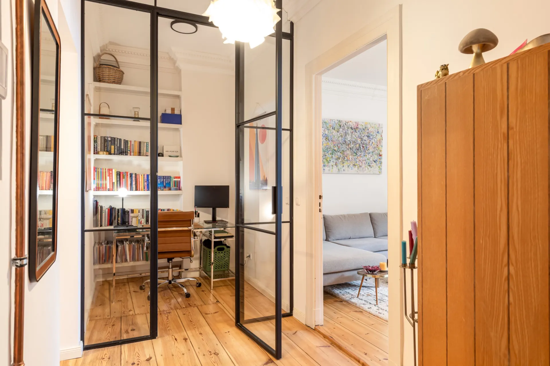 Kleines Homeoffice mit Glastüren, Bücherregal, Schreibtisch und Blick ins Wohnzimmer mit Sofa.