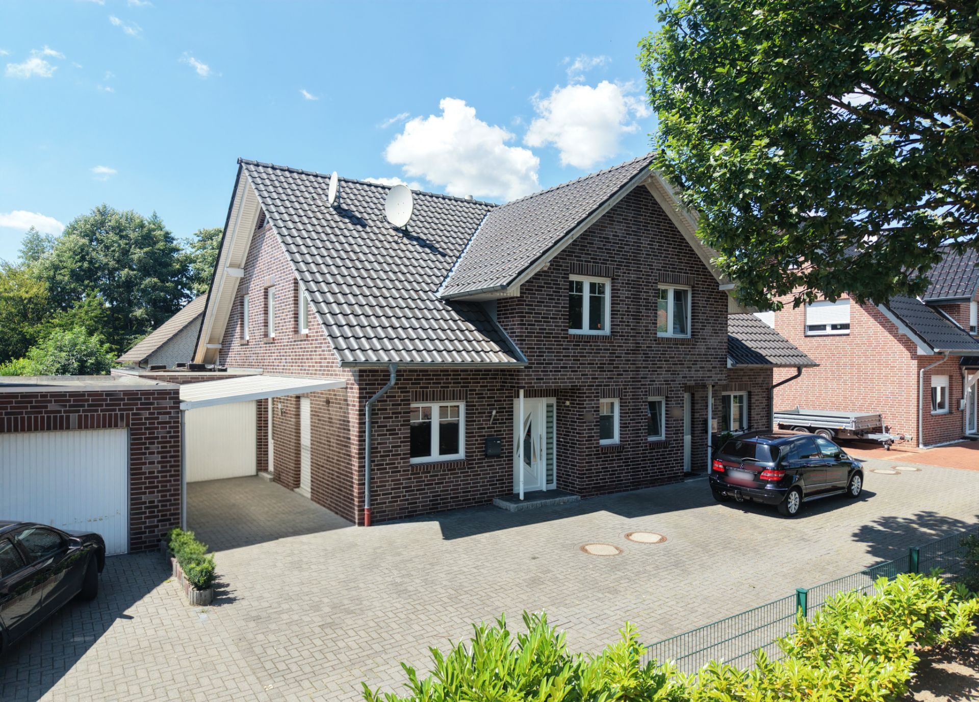 Zweistöckiges Backsteinhaus mit Garage, gepflastertem Hof und geparkten Autos bei Sonnenschein.