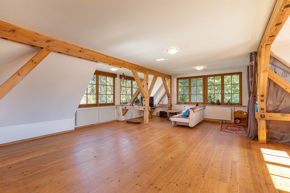 Großer heller Raum mit Holzboden, sichtbaren Holzbalken und Sofa unter mehreren Fenstern.