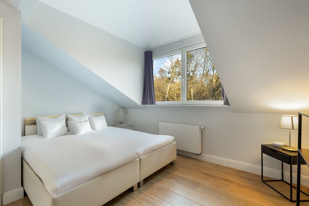 Helles Schlafzimmer mit Doppelbett, Fenster und Nachttisch mit Lampe unter Dachschräge.