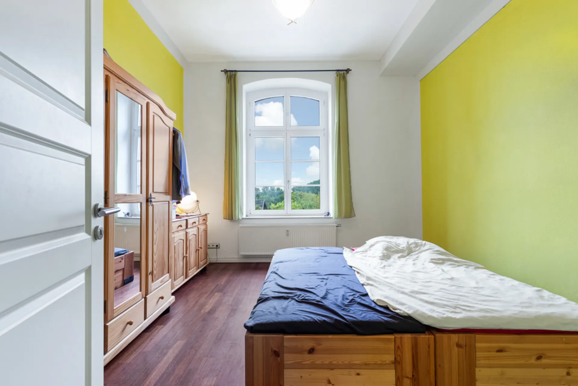 Helles Schlafzimmer mit Holzbett, Holzschrank und großem Fenster mit Blick nach draußen.