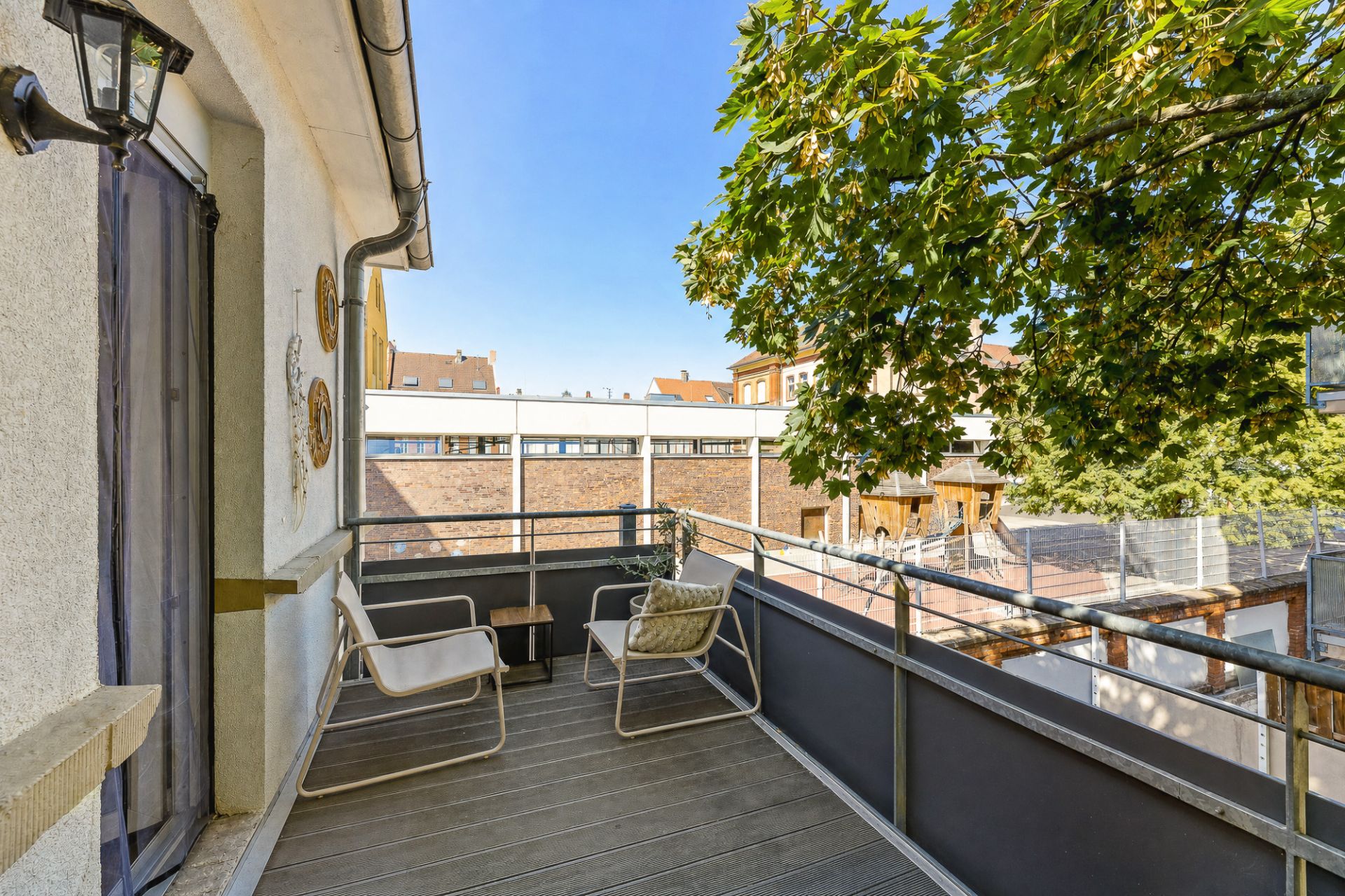 Balkon mit zwei Stühlen, kleinem Tisch und Blick auf Bäume und Nachbargebäude.