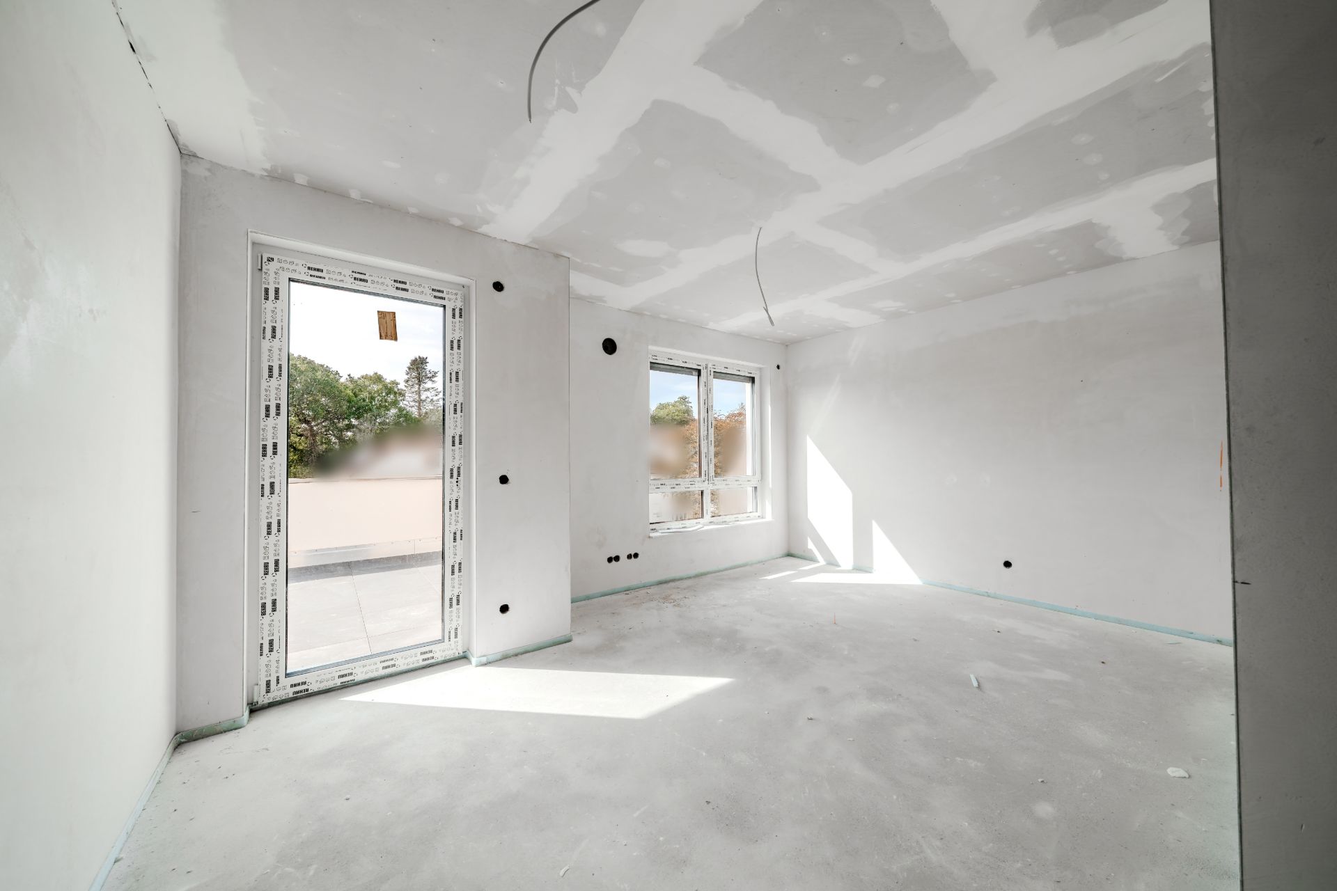 Helles Zimmer im Rohbau mit großen Fenstern und unvollständigem Boden.
