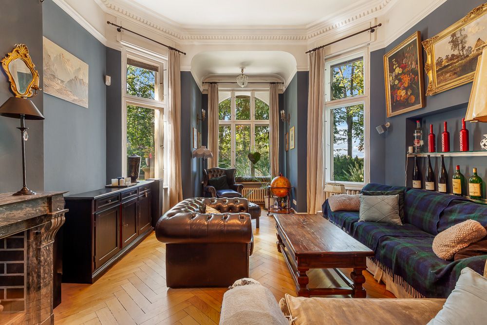 Wohnzimmer mit großen Fenstern, blauen Wänden, Ledersessel, Sofa und Holztisch.