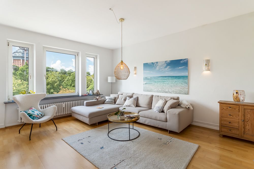 Helles Wohnzimmer mit Ecksofa, Sessel, Holzboden, Teppich und großen Fenstern mit Ausblick auf Bäume.