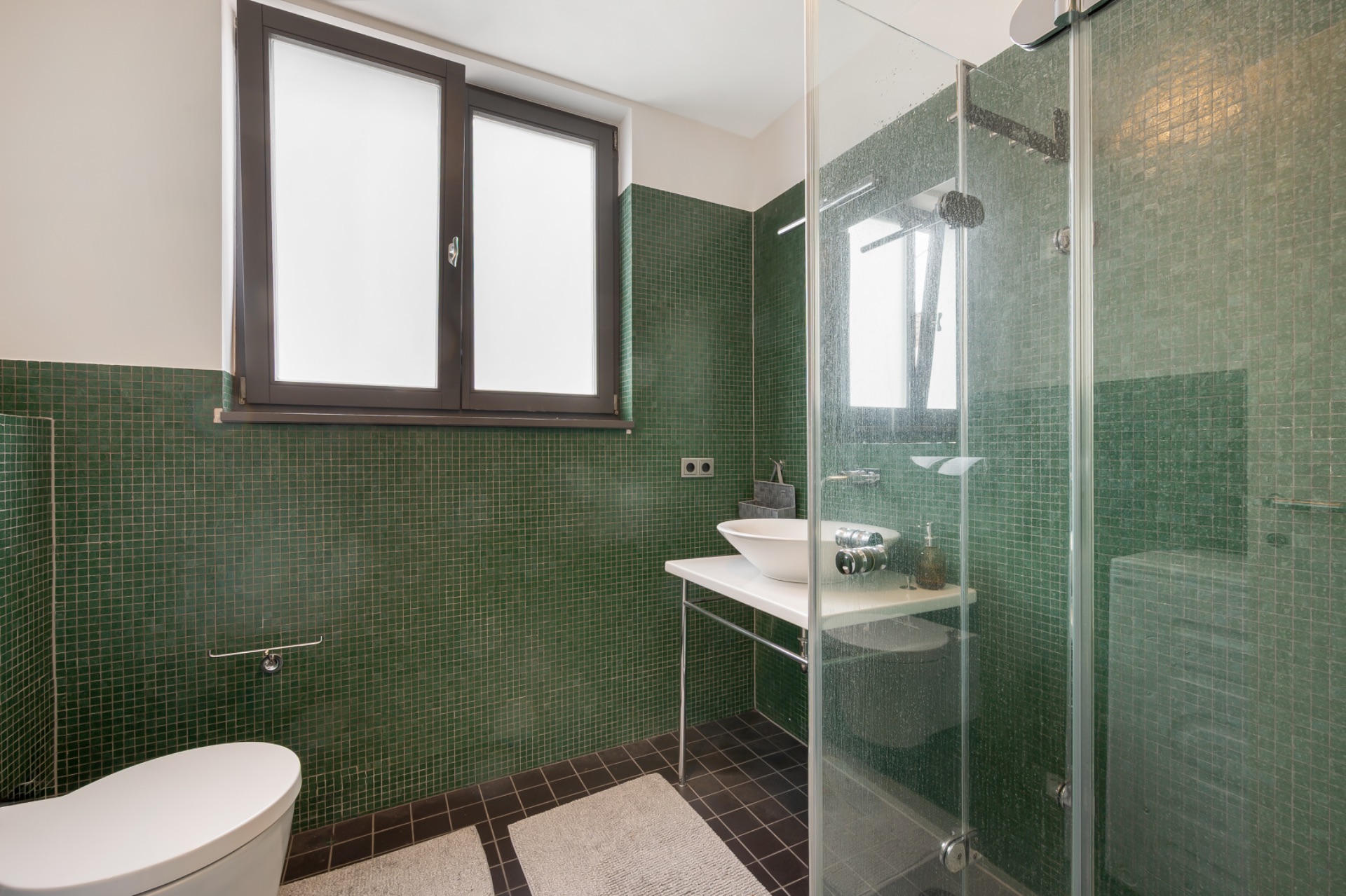Modernes Bad mit grünen Fliesen, WC, Waschtisch und verglaster Dusche.