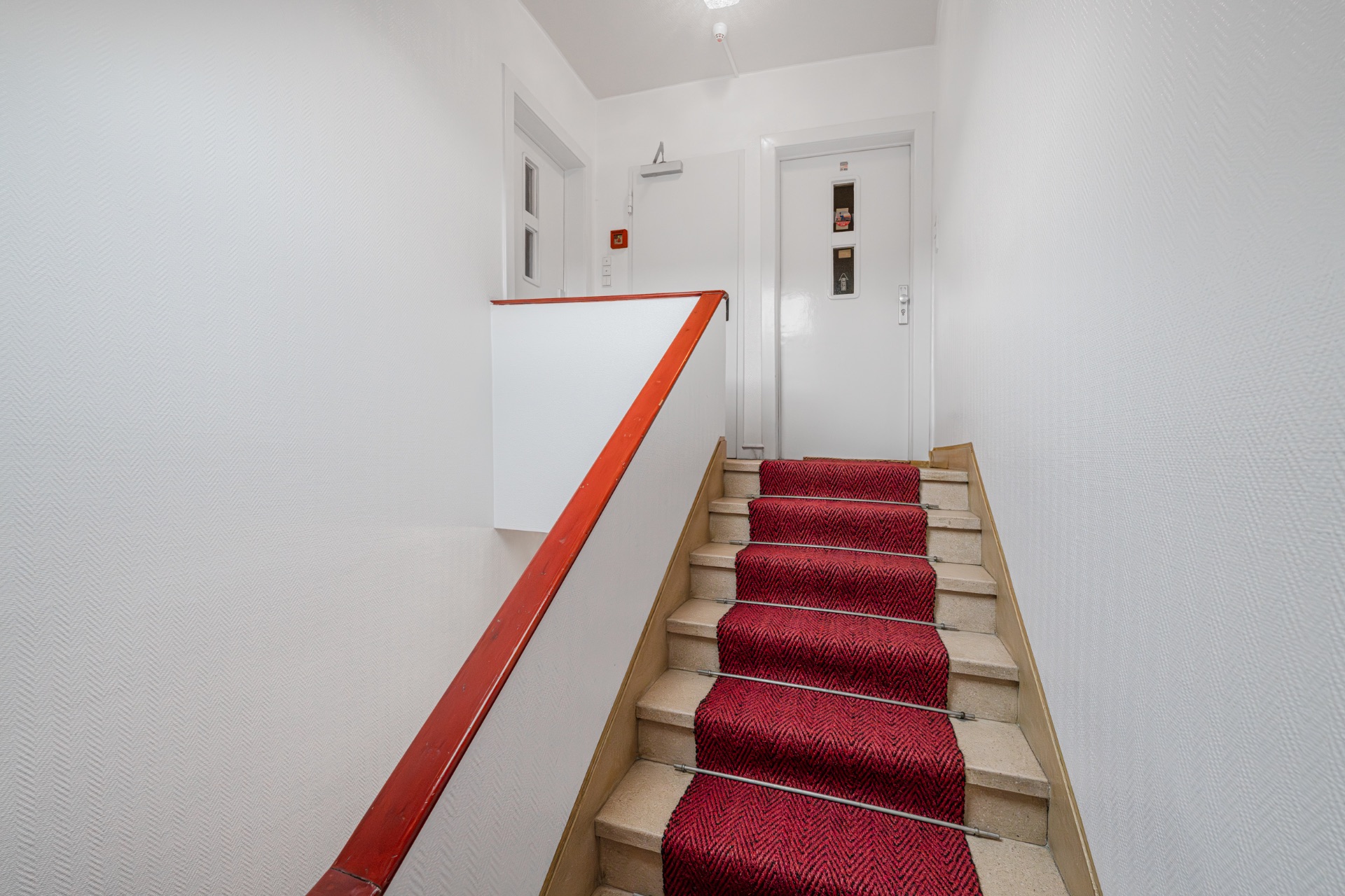 Treppenaufgang mit rotem Teppichläufer und weißen Wänden in einem Wohnhaus.