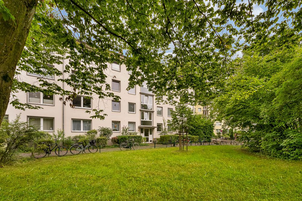 Mehrfamilienhaus mit Vorgarten, Bäumen und Fahrrädern am Gehweg.