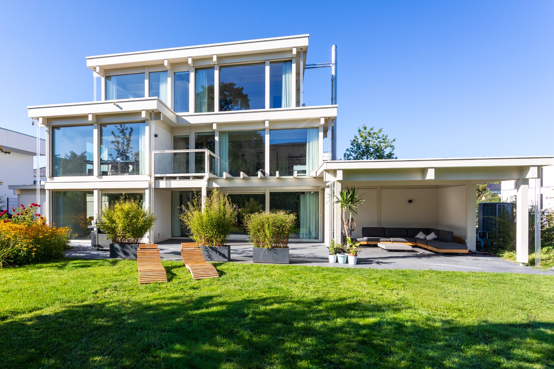 Modernes, dreistöckiges Haus mit großen Glasfenstern, Garten und Terrasse mit Liegestühlen.