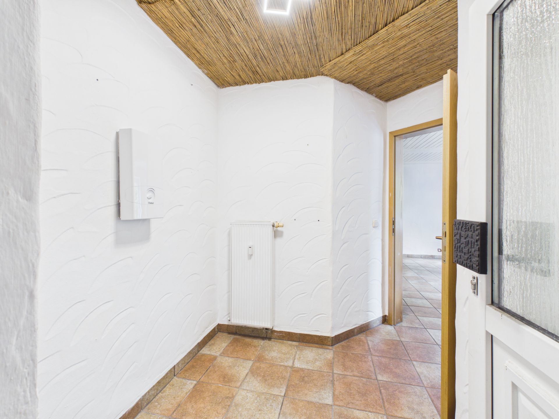 Heller Flur mit Fliesenboden, weißer Wand, Heizkörper und Holztür, Decke mit Holzverkleidung.