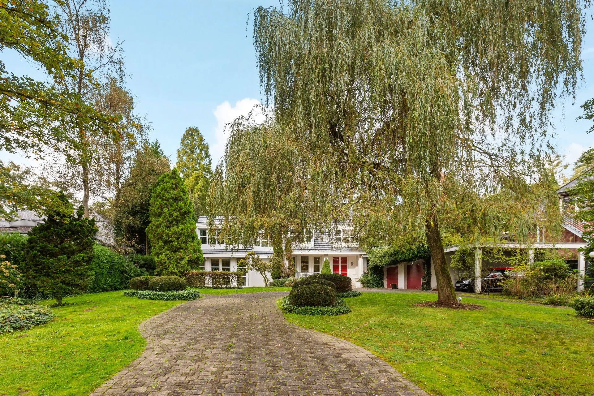 Großes Wohnhaus mit weißer Fassade, Auffahrt und gepflegtem Garten mit Baum im Vordergrund.
