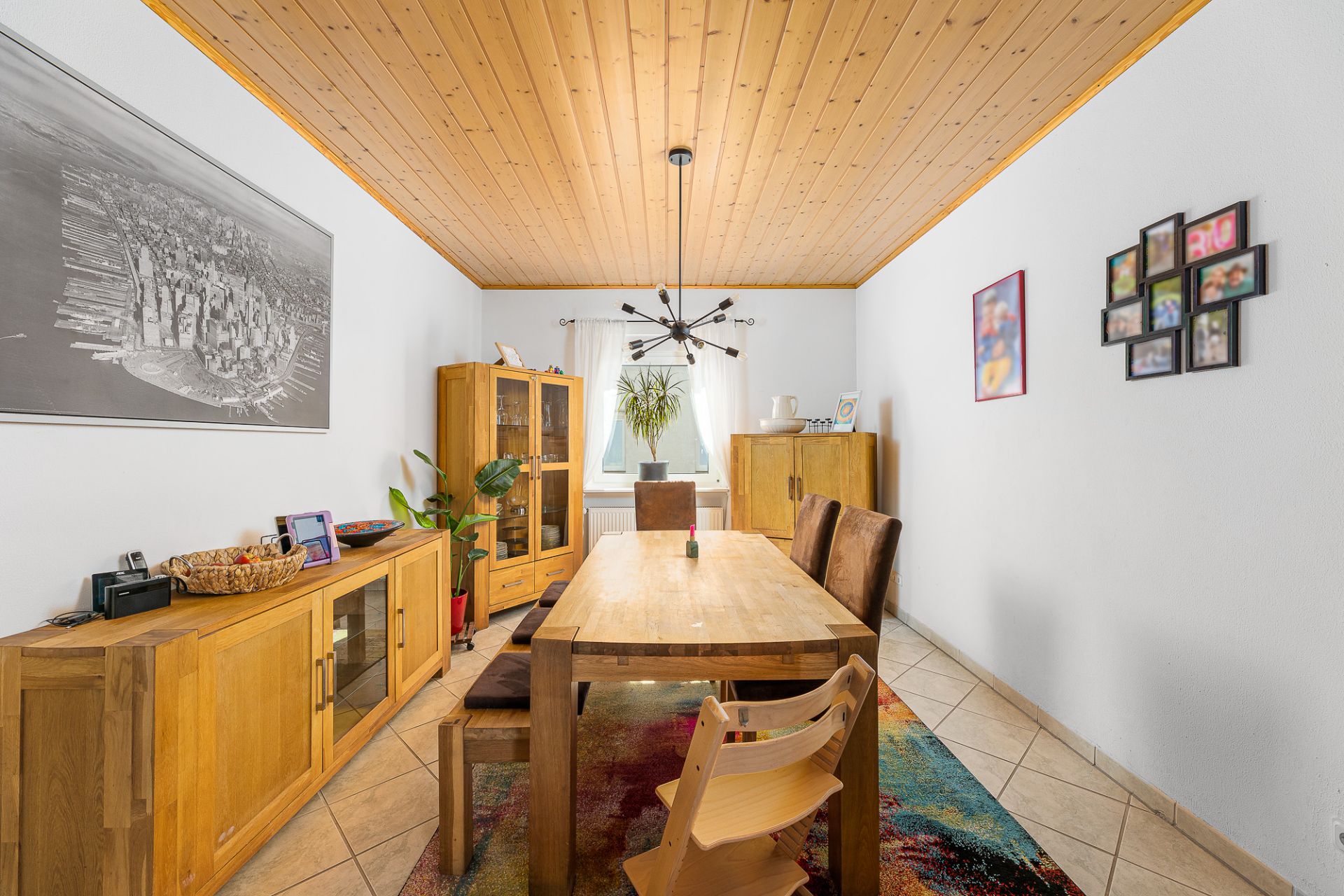 Esszimmer mit Holztisch, Stühlen, Holzdecke und buntem Teppich, hell durch Fenster mit Pflanze.