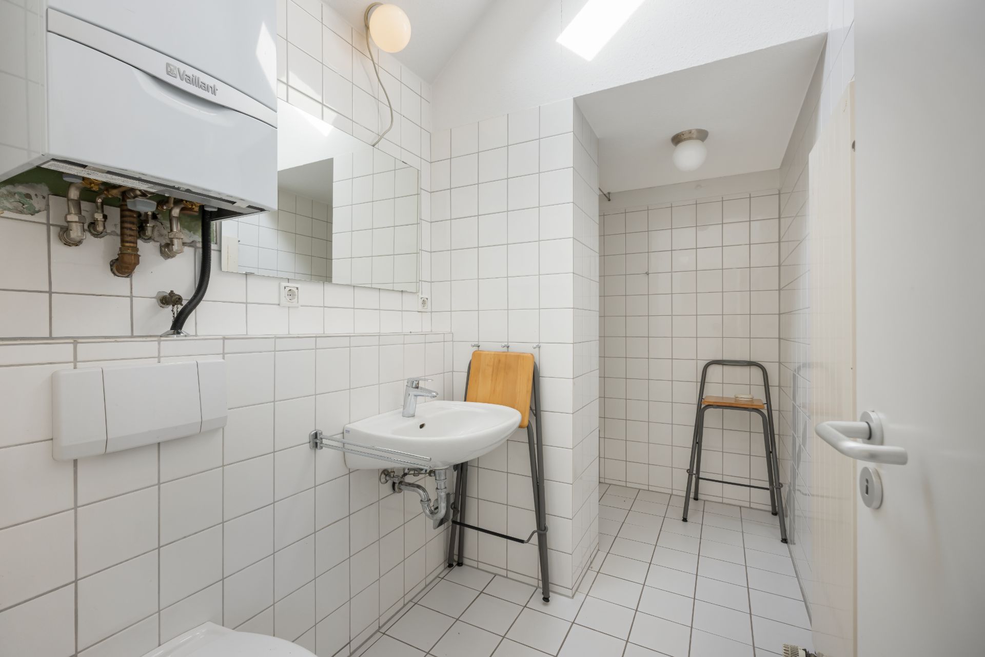 Helles Badezimmer mit weißen Fliesen, Waschbecken, Spiegel, Hocker und Wandheizung.