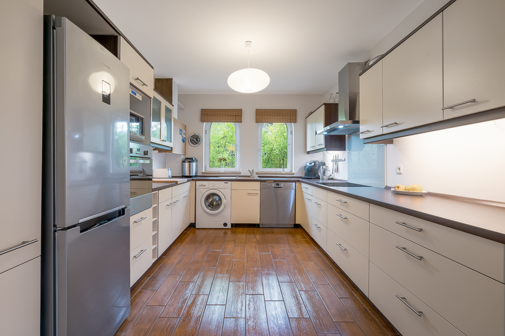 Moderne Küche mit weißen Schränken, Holzfußboden, Geräten und zwei Fenstern mit Blick ins Grüne.