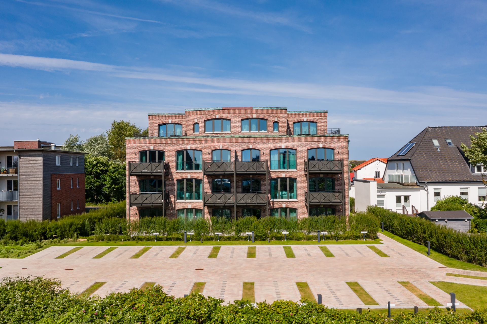 Backsteinwohnhaus mit Balkonen, davor leerer gepflasterter Parkplatz und grüne Hecken.