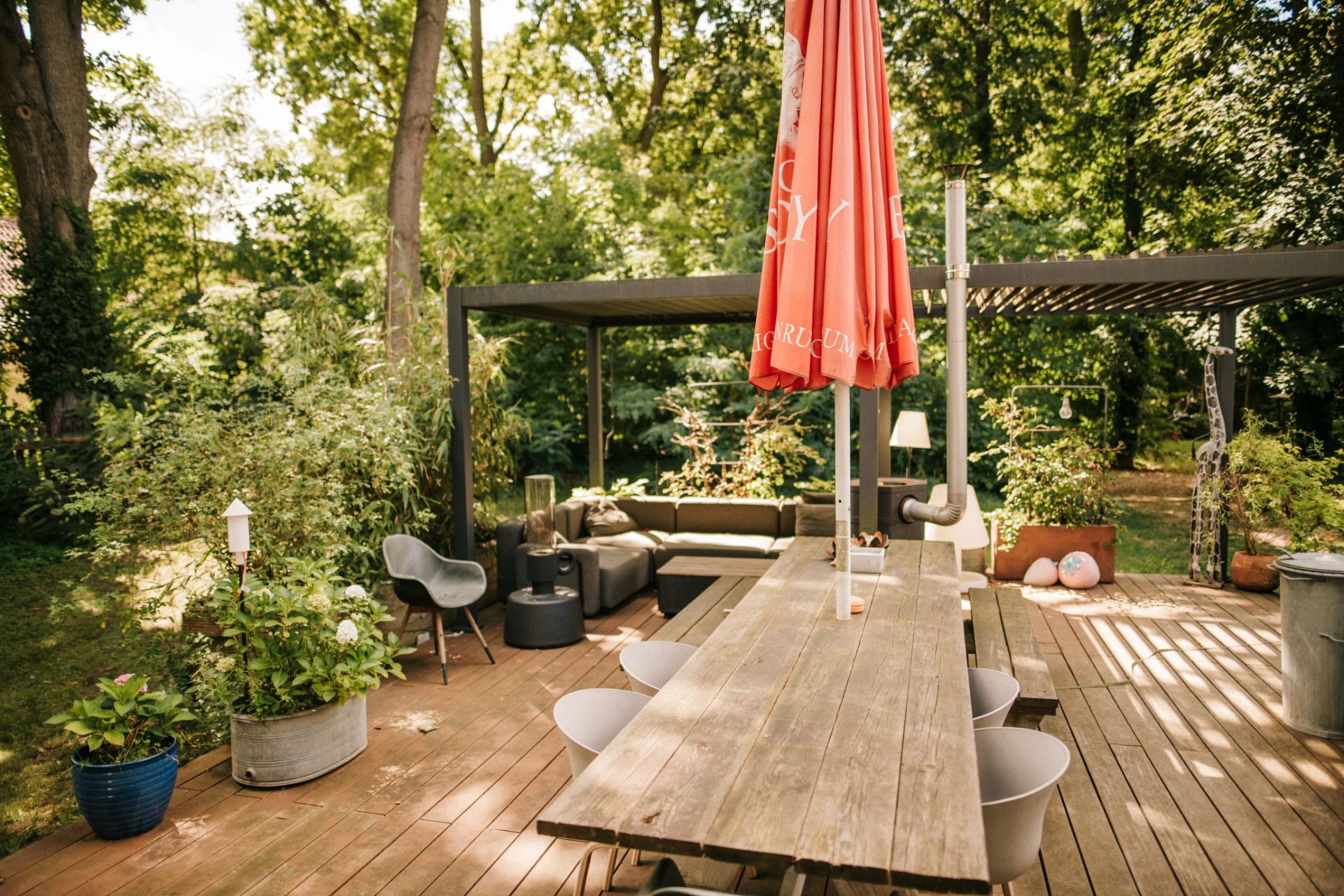 Große Holzterrasse mit langem Tisch, Stühlen, rotem Sonnenschirm und Sitzbereich im Garten.