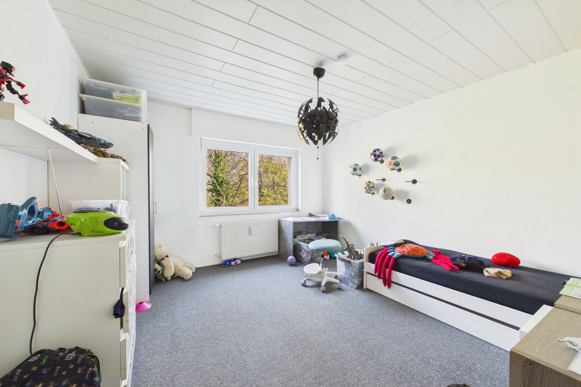 Helles Kinderzimmer mit Einzelbett, Spielzeug, Regal, Schreibtisch und Fenster mit Blick ins Grüne.