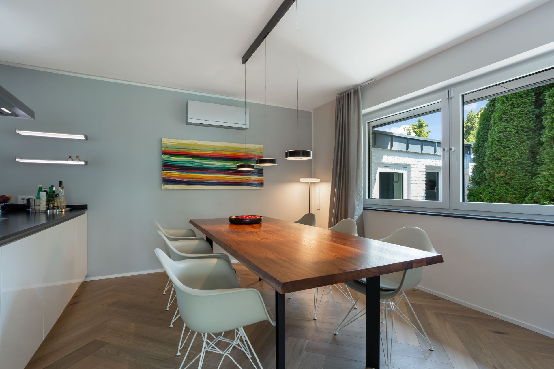 Esszimmer mit Holztisch, sechs weißen Stühlen, großem Fenster und buntem Bild an der Wand.