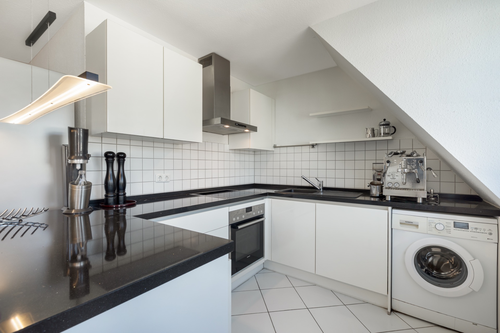 Moderne weiße Küche mit Waschmaschine, Kaffeemaschine und schwarzer Arbeitsplatte.