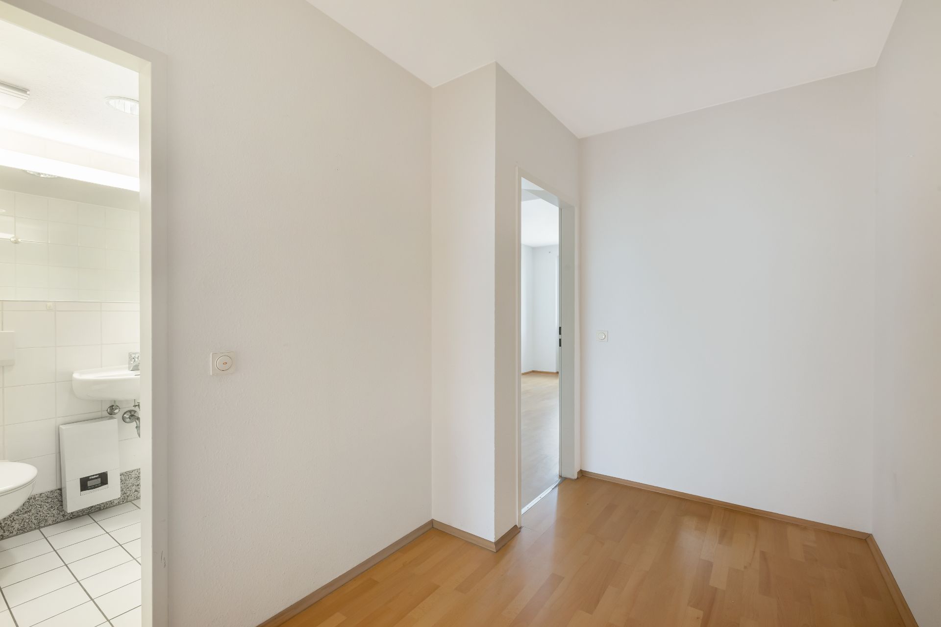 Kleiner Flur mit Holzboden, Blick ins Badezimmer mit weißen Fliesen und Waschbecken.