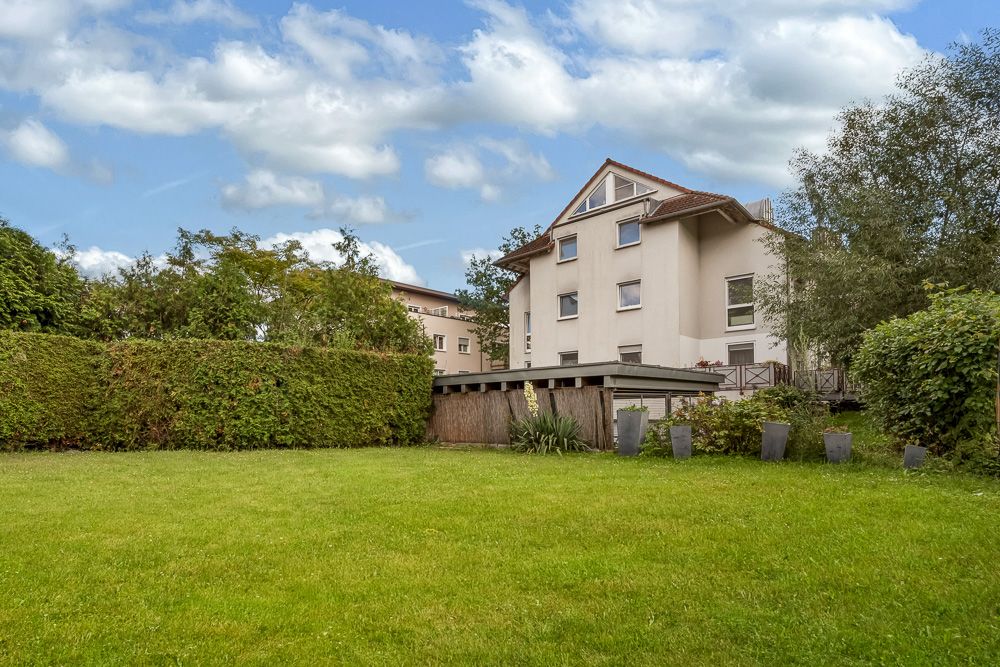 Mehrfamilienhaus mit Garten, Hecke und Bäumen im Vordergrund, Himmel mit Wolken.